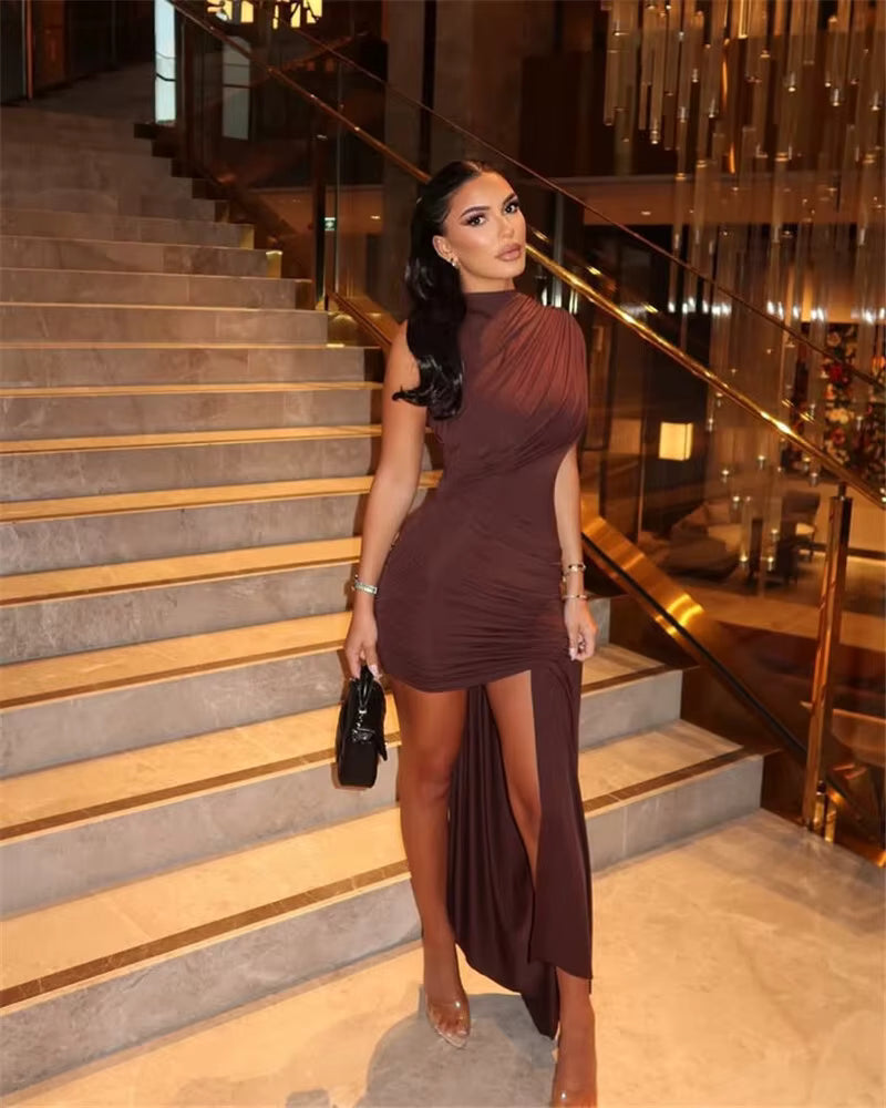 Elegant woman in a brown one-shoulder draped mini dress with a long side train, posing on a gold-lit staircase in a upscale venue