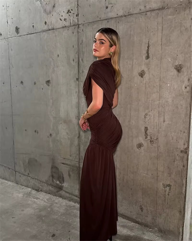 Model in a fitted chocolate brown asymmetric mini dress with a dramatic side sash, standing against a textured concrete wall